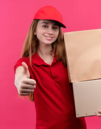 smiling-young-delivery-girl-red-uniform-holding-boxes-showing-thumb-up-isolated-pink-space-with-copy-space